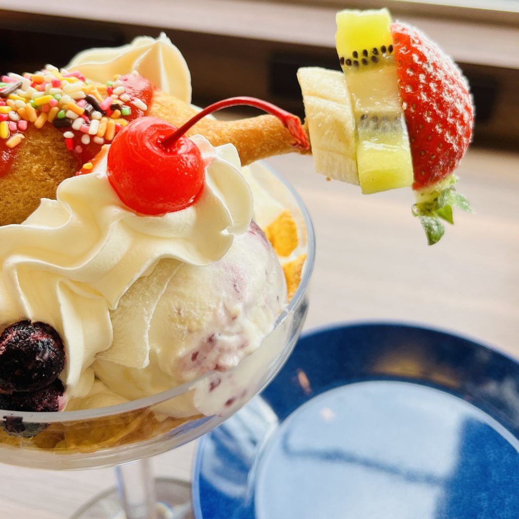 魅惑のアメリカンドッグパフェのアップ。串に刺さったイチゴやキウイ、バナナがアメリカンドッグの横に並ぶ
